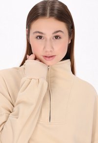 Beige zip-up sweatshirt med hög krage, ribbade mudd och mjuk textur. Modellen håller tyget nära ansiktet, vilket visar plaggets avslappnade passform.