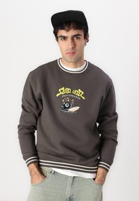 Felpa grigia con logo ricamato, caratterizzata da accenti gialli e rossi, scollatura rotonda con dettaglio a righe e vestibilità rilassata. Cappello nero.
