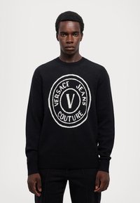 Črn pleten pulover z velikim belim logotipom, ki vsebuje besedilo "VERSACE JEANS COUTURE" in črko "V" na sredini. Rebrasti manšetni.