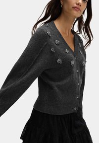 Szary kardigan z prążkowanym wykończeniem, zdobiony kwiatowymi aplikacjami i zapinany na guziki, w połączeniu z czarną warstwową spódnicą.