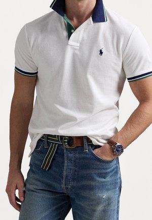 Weißes Polohemd mit einem marineblauen und grünen Kragen, kombiniert mit blauen Jeans. Verfügt über kontrastierende Streifen an den Ärmeln und ein kleines Logo auf der Brust.