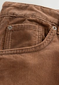 Bruin corduroy stof met verticale ribbels, voorzien van een voorkantzak met metalen klinknageldetails. De textuur lijkt zacht en duurzaam.