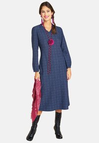 Marineblaues Langarmkleid mit subtilen Mustern, V-Ausschnitt und taillierter Taille. Kombiniert mit einem roten Bandana und schwarzen Schnürstiefeln.