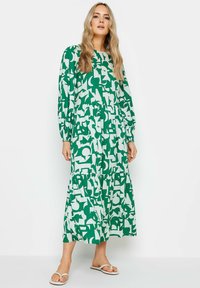 Langes grünes Kleid mit weißen abstrakten Mustern, featuring lange Ärmel, einen gestuften Rock und einen elastischen Bund. getragen mit weißen Sandalen.