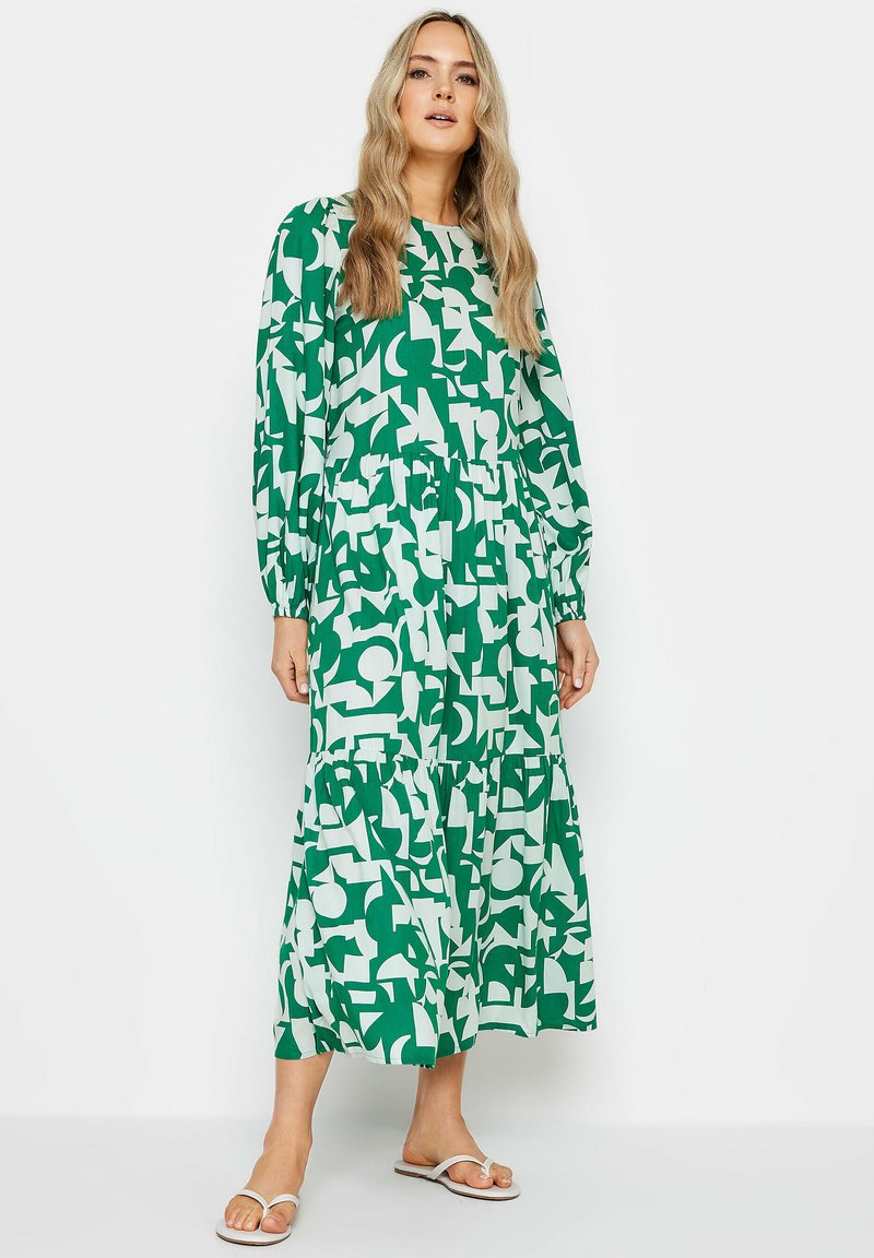 Langes grünes Kleid mit weißen abstrakten Mustern, featuring lange Ärmel, einen gestuften Rock und einen elastischen Bund. getragen mit weißen Sandalen.