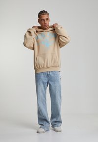 Felpa beige con testo grafico blu e design di leone, abbinata a jeans a gamba larga azzurri e sneakers bianche. Tessuto liscio, vestibilità casual.
