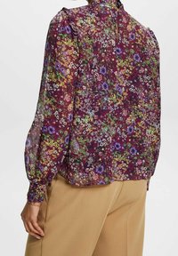Kwiecista bluzka wykonana z przezroczystej tkaniny o bordowym tle z wielokolorowymi wzorami kwiatowymi. Szczegóły obejmują bufiaste rękawy i zapinany na guziki tył.