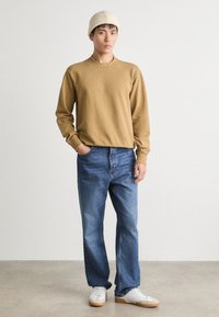 Sudadera beige, jeans azules y zapatillas blancas con suelas marrones; el modelo lleva un gorro de punto beige; ajuste casual. Textura suave y diseño sencillo.