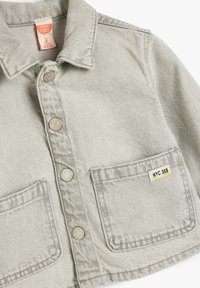 Lichtgrijze denim babyjas met een kraag, twee voorzakken en vijf drukknoopsluitingen. Bevat een "NYC SK8" label op de rechterzak.