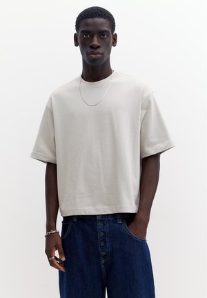 Giovane uomo con indosso una maglietta beige larga, jeans blu scuro con bottoni e collana e bracciale d'argento, in piedi davanti a uno sfondo bianco.