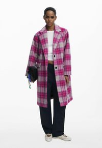Cappotto scozzese nei colori rosa e grigio, realizzato in tessuto testurizzato, con un design doppiopetto e due tasche frontali. Indossato sopra una maglietta bianca.