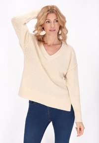 Pulover tricotat de culoare crem, cu croi lejer, de tip V, cu tiv striat și model deschis. Asortat cu blugi de culoare bleu închis.