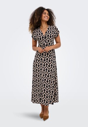 Donna con capelli ricci che indossa un vestito midi nero e beige a fantasia e ballerine marroni con punta, sorridente e con le mani in vita.