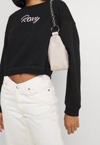Persona con felpa nera corta con il testo "Roxy", jeans bianchi a vita alta e una piccola borsa a spalla beige con catena.