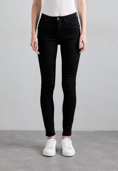 Jean skinny en denim noir avec cinq poches, fermeture à bouton et passants de ceinture, associés à des baskets blanches sur un sol gris.