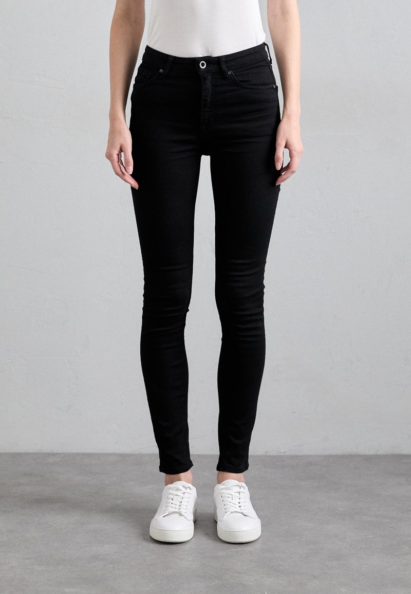 Schwarze Skinny-Jeans aus Denim mit fünf Taschen, Knopfverschluss und Gürtelschlaufen, kombiniert mit weißen Turnschuhen auf einem grauen Boden.