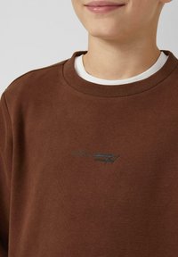 Brązowa bluza z prążkowanym okrągłym dekoltem i białym wewnętrznym kołnierzem. Delikatne zielone hafty z napisem "ARRIVAL crafted." Miękka faktura.