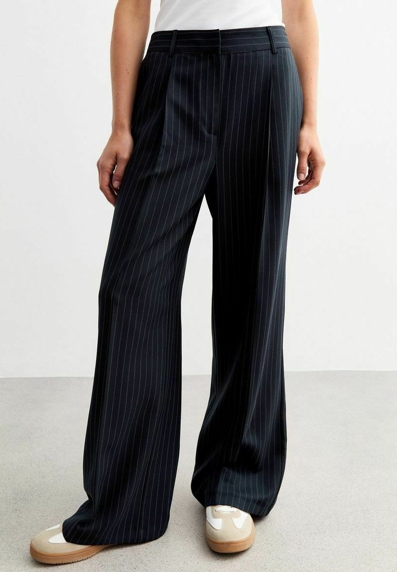 Personne portant un pantalon large noir à fines rayures, associé à des baskets blanches et beiges, debout sur un sol gris contre un mur blanc.