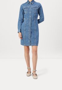 Abito camicia in denim azzurro chiaro con maniche lunghe, chiusura frontale con bottoni, due tasche al petto e borchie decorative sulla zona delle spalle.