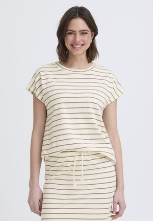 Smilende kvinde med kort brunt hår iført et cremefarvet sæt med tynde brune vandrette striber og en nederdel med snøre.