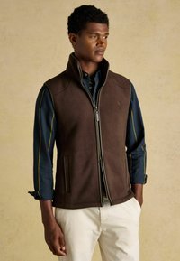 Hnedý flísový vest s vysokým golierom, predným zipsom a bočnými vreckami. Nosený cez modrú košeľu s zelenými a žltými pruhmi.