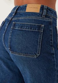 Tunna blå denimjeans med medelhög midja, med en enkel bakficka, orange sömmar och en märkt tan läderlapp vid midjan.