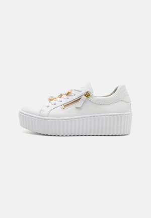 White sneaker with a textured rubber platform sole, gold hardware accents, decorative zippers, and lace-up front. Smooth leather material.