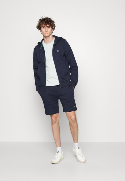 Un sweat à capuche bleu marine avec une fermeture éclair frontale et des cordons, associé à un short assorti. Des baskets blanches complètent la tenue. Un accent vert sur le sweat à capuche.