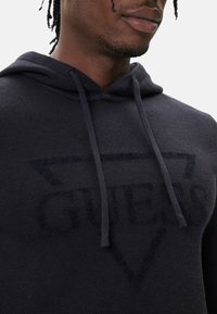 Felpa con cappuccio blu navy scuro in tessuto morbido con logo "GUESS" tono su tono e coulisse. Il design include un contorno del logo triangolare.