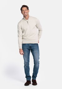 Beige zip-up trui van een zachte stof, gecombineerd met blauwe jeans en donkerbruine laarzen. De trui heeft een geribde zoom en manchetten.