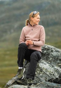 Jasnoróżowa polarowa kurtka z zamkiem, czarne spodnie i zielone buty sportowe. Siedząca na głazie w naturalnym otoczeniu na świeżym powietrzu.