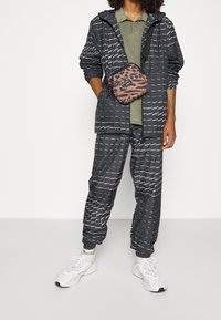 Černá dešťová bunda a kalhoty s bílým textovým vzorem, spárované se zeleným polo tričkem a hnědým batohem s leopardím potiskem. Bílé tenisky.