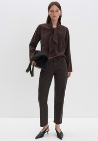 Donna che indossa una blusa marrone scuro con cravatta, pantaloni abbinati, scarpe con tacco nero a slingback, mentre tiene una borsa nera piegata contro uno sfondo bianco.