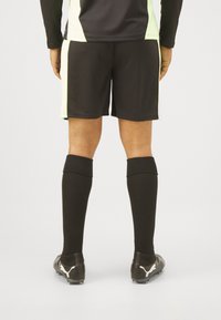 Schwarze Sportshorts mit limettengrünen Akzenten, kombiniert mit schwarzen Schienbeinschonern und schwarzen Fußballschuhen mit einem weißen Logo.
