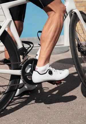 Zapato de ciclismo blanco con un diseño elegante, parte superior perforada y acentos negros, asegurado a un pedal de bicicleta. Se ve la pierna de un ciclista, mostrando un detalle muscular.