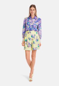 Langärmlige Bluse mit floralem Muster in Lila und Grün; kombiniert mit hochgeschnittenen gelben Shorts, die ein ähnliches Blumenmuster aufweisen.