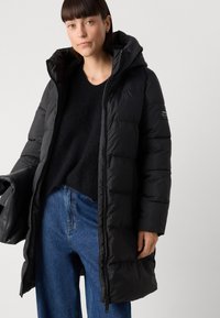 Black puffer coat with a hood, featuring quilted panels, zip closure, and a label on the sleeve. Paired with a black sweater and blue jeans.