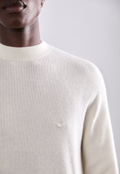 Pulover ribbed de culoare crem, cu guler înalt și broderie subtilă a logo-ului pe piept. Textură netedă cu dungi verticale.
