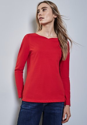 Rotes Langarmshirt mit Rundhalsausschnitt, aus weichem Baumwollstoff. Kombiniert mit dunkelblauen Jeans. Schlichtes und figurschmeichelndes Design.