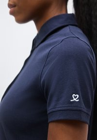 Γυναίκα που φοράει μπλε σκούρο πόλο με ένα μικρό λευκό λογότυπο σε σχήμα καρδιάς στο μανίκι, εμφανιζόμενη από το πλάι και από τον ώμο και πάνω.