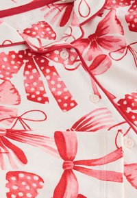 Rotes und pinkes gemustertes Stoff mit Schleifen und Polka-Dots auf weißem Hintergrund. Kurzarm-Design mit Knöpfen und Taschendetail.