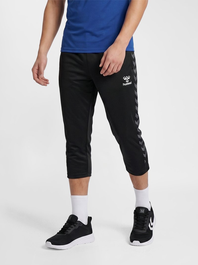 Persona que lleva pantalones deportivos negros hasta la mitad de la pantorrilla con patrón en forma de cheurón lateral, calcetines blancos, zapatillas negras y una camisa azul, de pie.