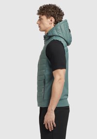 Viegls, polsterēts veste teāl krāsā ar kapuci, ar iešūtu kabatu, elastīgu apakšmali un solid black piedurknēm krekli zem tā.
