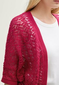 Cardigan en tricot ouvert fuchsia avec un motif texturé et des poignets côtelés, superposé à un top blanc ajusté. La conception présente une coupe décontractée.