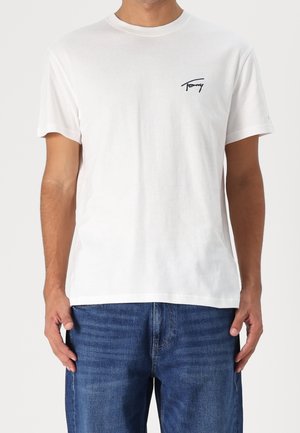 Homme portant un T-shirt blanc à manches courtes avec un petit texte noir "Tommy" sur le côté gauche de la poitrine, associé à un jean bleu.