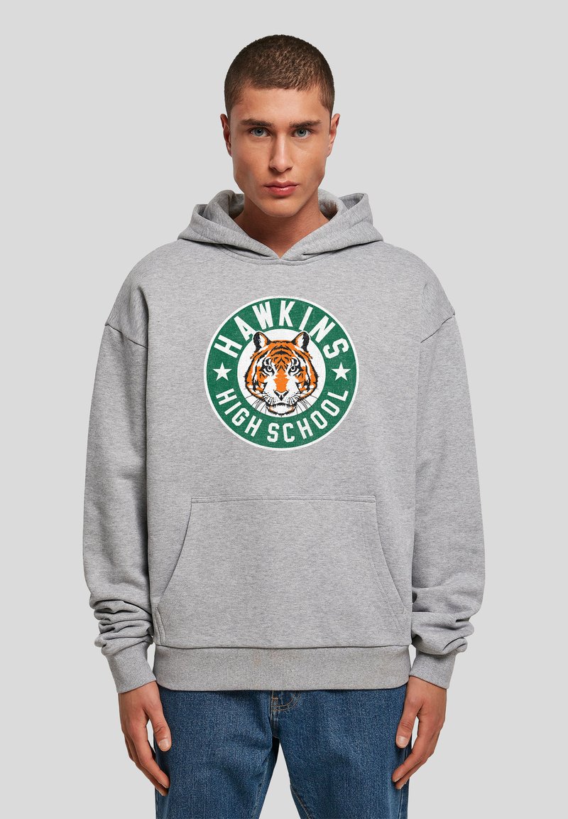 Sweat-shirt gris avec un grand logo circulaire de tigre, texte "HAWKINS HIGH SCHOOL" en vert et blanc. Comprend une poche avant et une capuche.
