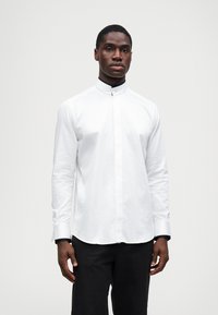 Hvit langermet skjorte med høy krage, har en jevn tekstur, subtil glans og kontrasterende svarte mansjetter.