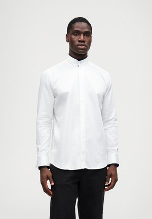 Chemise blanche à manches longues avec un col haut, présentant une texture lisse, un léger éclat et des poignets noirs contrastants.