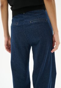 Dunkelblaue Denim-Hose mit lockerer Passform, zwei Gesäßtaschen und glattem Stoff, sichtbare Nähte entlang des Taillenbunds.