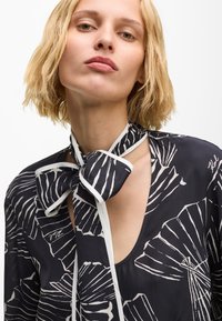 Fekete blúz fehér levélmintával, nyakkendős részlettel, V-nyakkivágással. Könnyű anyag, sima textúra, kontrasztos szegély. Minimalista dizájn.